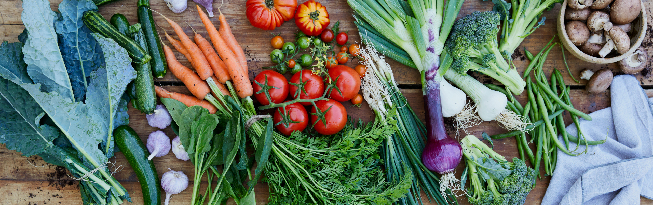 Overhead shot of a table full of fresh kale, cucumber, carrots, tomatoes, broccoli, mushrooms and green beans.
